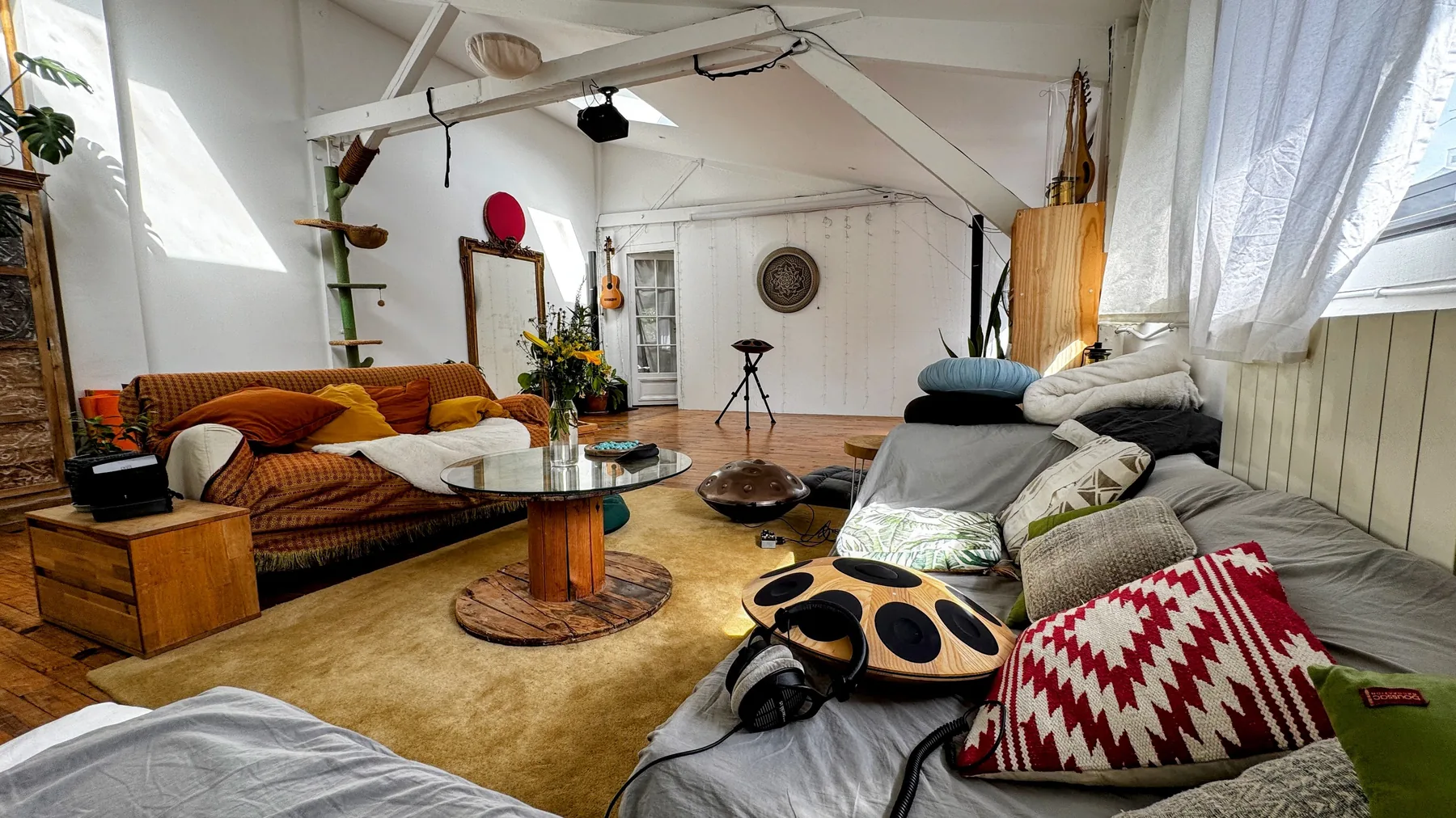 Le coin salon du showroom : un handpan acoustique posé au sol près de la table basse, un handpan électronique Neotone en bois avec son casque sur les coussins, un grand canapé orange, un bouquet de fleurs et le mur de guirlandes lumineuses au fond.